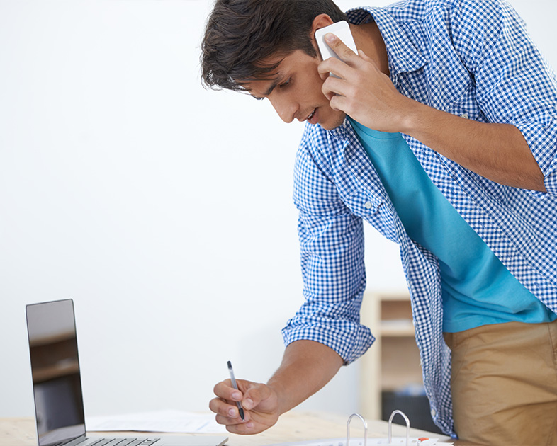Hombre joven con camisa a cuadros, hablando por teléfono mientras escribe en una hoja frente a una computadora portátil. El entorno es luminoso y minimalista.