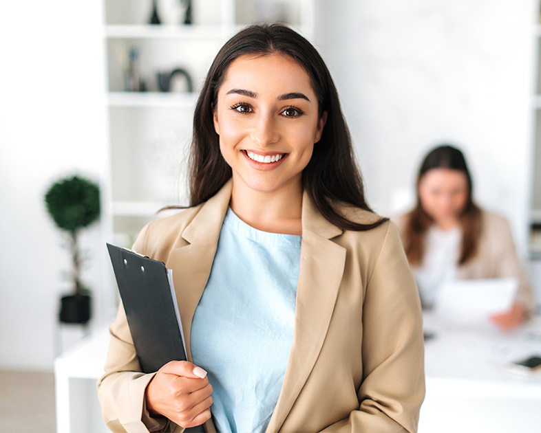 Mujer sonriente con blusa azul y chaqueta beige sostiene una carpeta, en un entorno de oficina con otra persona al fondo.