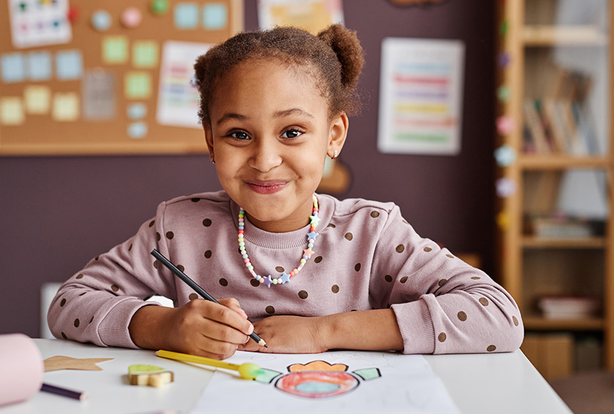 Niña sonriente con cabello rizado, usando un suéter con puntos, dibujando en una mesa. Fondo con un tablero y estanterías. Colores cálidos predominan en el entorno.