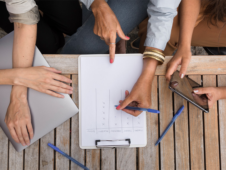Manos de varias personas sobre una mesa de madera, una persona escribe en una hoja con gráficos, mientras otras señalan y sostienen dispositivos móviles. Predominan tonos neutros.