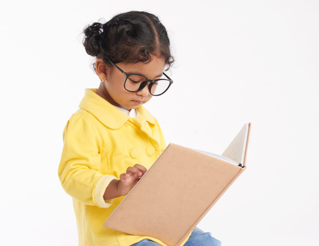Niña con gafas, vestida con un abrigo amarillo, lee un libro en un fondo blanco.