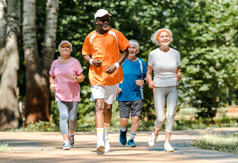 Cuatro personas mayores corriendo en un sendero, con árboles verdes de fondo. Visten ropa deportiva de diferentes colores.