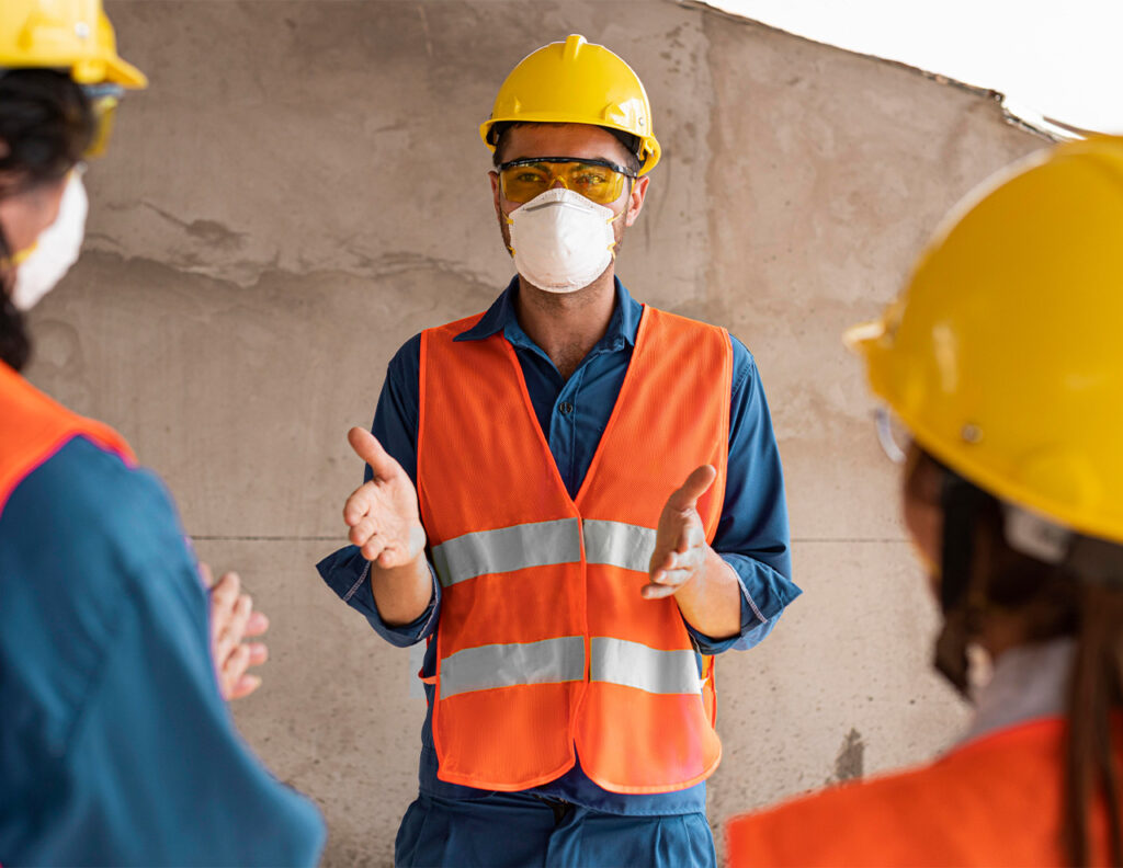Trabajador con casco y chaleco reflectante explica a otros compañeros en un entorno de construcción, todos usando mascarillas.