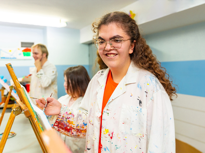 Tres personas en un taller de arte, una sonríe con un pincel, mientras las otras pintan en el fondo.