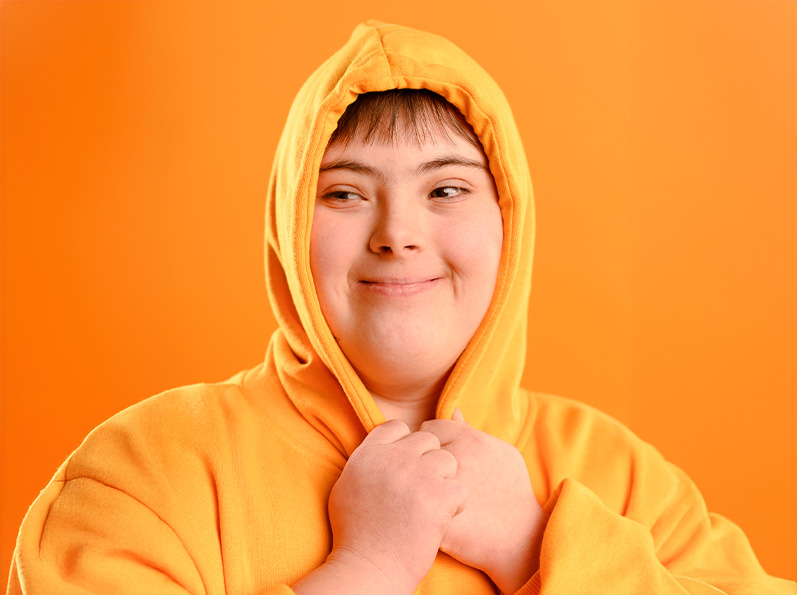 Niño con sudadera naranja, sonriendo, en un fondo naranja. Manos juntas cerca del pecho, expresión amigable.
