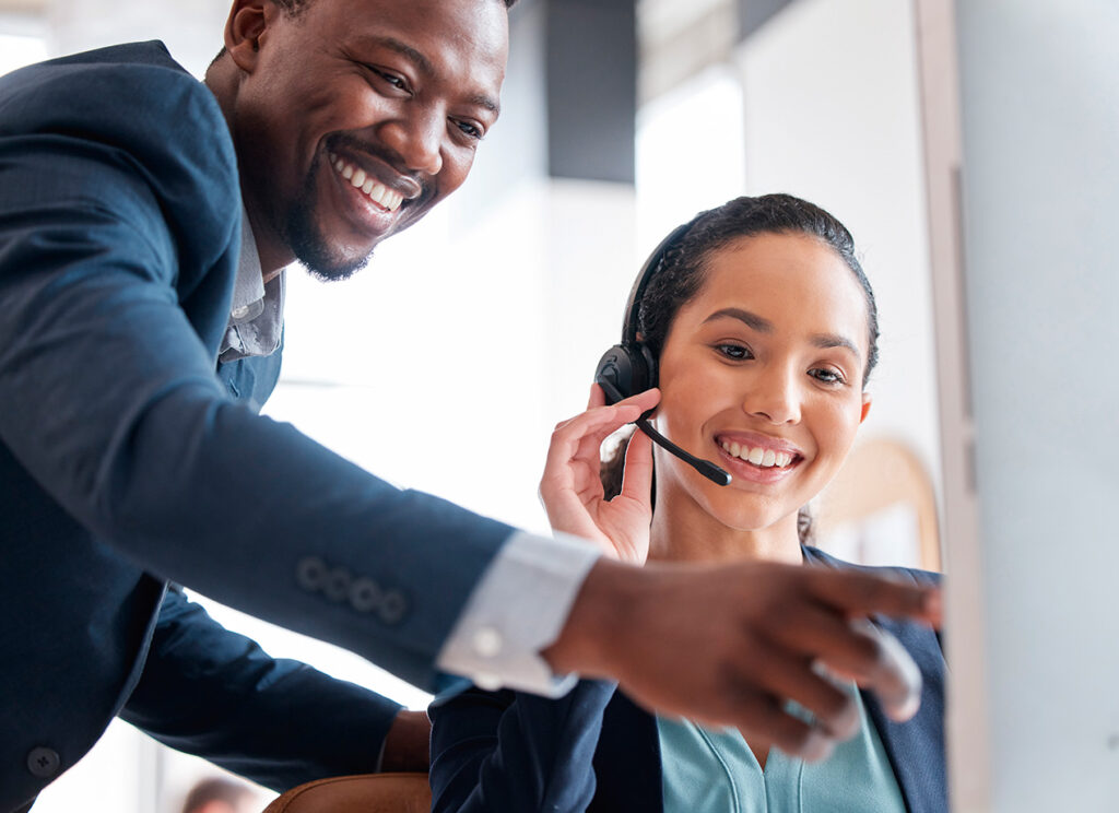 Dos personas en un entorno de oficina, una mujer con auriculares sonriente y un hombre señalando hacia una pantalla. Predominan colores neutros y un ambiente laboral.