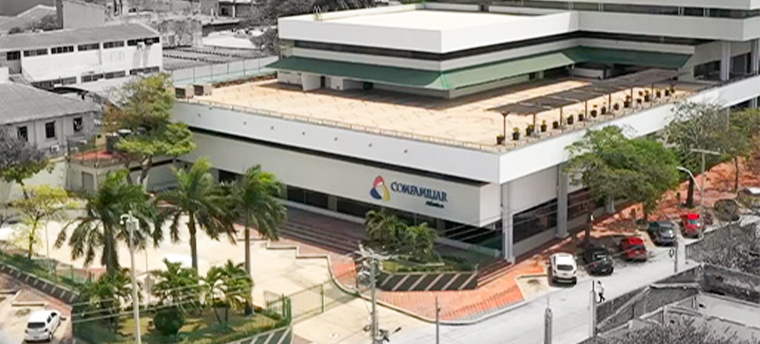Edificio moderno con el logo de Comfamiliar, rodeado de áreas verdes y estacionamiento. Predominan tonos claros y un entorno urbano con vehículos y árboles.