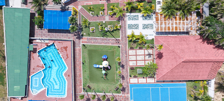 Zona recreativa al aire libre con piscina, áreas verdes, juegos infantiles y canchas de tenis. Predominan colores azul, verde y terracota. Se observan palmeras y personas disfrutando.