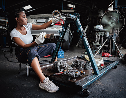 Mujer sentada en un taller mecánico, sosteniendo una pieza de motor con guantes. A su lado, herramientas y maquinaria en un entorno industrial. Predominan colores oscuros y metálicos.