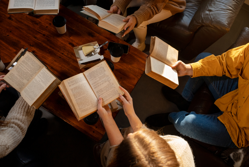 Varias personas sostienen libros abiertos alrededor de una mesa de madera. Se observan tazas de café y un trozo de pastel. La iluminación es cálida y suave.