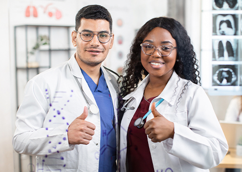 Dos profesionales de la salud, un hombre y una mujer, sonríen y hacen un gesto de pulgar arriba. Ambos visten batas blancas y gafas, en un entorno clínico con gráficos médicos.