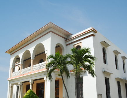 Casa de dos pisos con arcos en la fachada, rodeada de palmeras y un jardín pequeño, bajo un cielo despejado y soleado. Predomina el color blanco en las paredes.