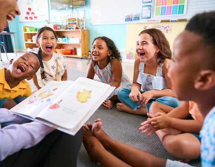 Grupo de niños sentados en el suelo, riendo y mirando un libro. Una persona adulta les lee, en un aula con colores vivos y materiales educativos visibles.