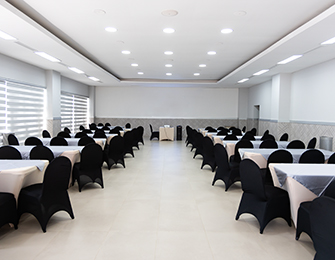 Salón amplio con mesas cubiertas, sillas negras y luz blanca. Las paredes son de color claro y hay ventanas con cortinas. Espacio preparado para eventos.