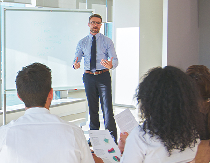 Hombre vestido formalmente presenta información frente a un grupo de personas sentadas. Se observa una pizarra blanca y documentos con gráficos en manos de los asistentes.