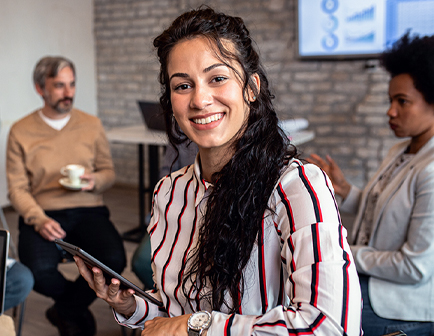Mujer sonriente con camisa de rayas, sosteniendo una tablet, en un entorno de oficina. Al fondo, dos personas conversan y hay pantallas con gráficos. Colores neutros predominan.