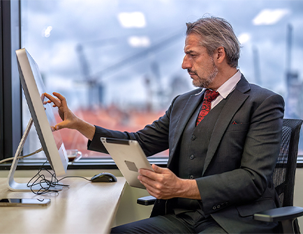Hombre de mediana edad, vestido con traje, interactuando con una computadora en un entorno de oficina. Sostiene una tablet mientras observa la pantalla. Fondo urbano con grúas visibles.