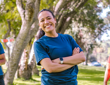 Mujer sonriente con camiseta azul, brazos cruzados, en un parque con árboles y otras personas al fondo. Ambiente natural y luminoso.