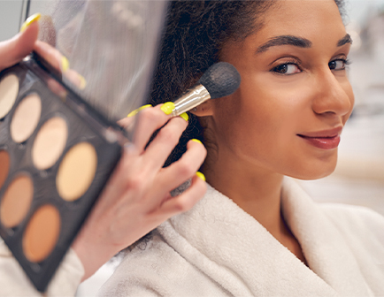 Mujer con piel morena sonríe mientras se le aplica maquillaje en las mejillas. Sostiene un albornoz blanco y hay una paleta de colores en primer plano.