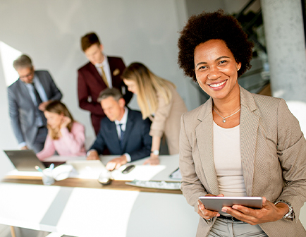 Mujer sonriente con tablet en mano, en primer plano, rodeada de personas en un entorno de oficina. Fondo con hombres y mujeres trabajando en mesas y computadoras. Colores claros y profesionales.