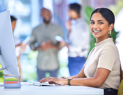 Mujer sonriente sentada frente a una computadora en un entorno de oficina moderno, con personas conversando al fondo y plantas decorativas. Predominan tonos claros y naturales.