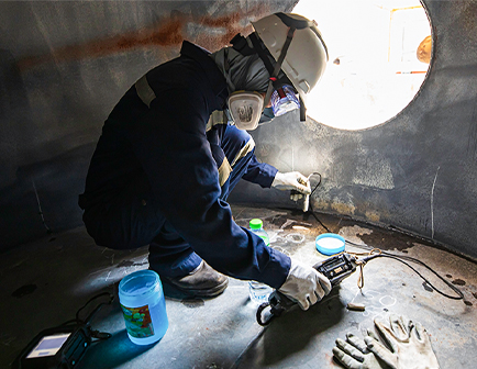 Trabajador en un espacio confinado, usando equipo de protección personal, mide con un dispositivo mientras está agachado. Se observan una botella de agua y guantes en el suelo.