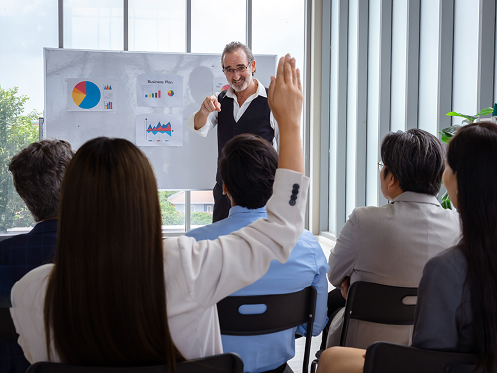 Hombre presentando en una sala con gráficos en una pizarra, mientras miembros de la audiencia levantan la mano para hacer preguntas. Ventanales permiten la entrada de luz natural.