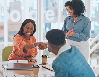 Dos mujeres y un hombre en una reunión. Una mujer sonriente estrecha la mano del hombre, mientras la otra mujer observa y aplaude. Hay tazas de café sobre la mesa.