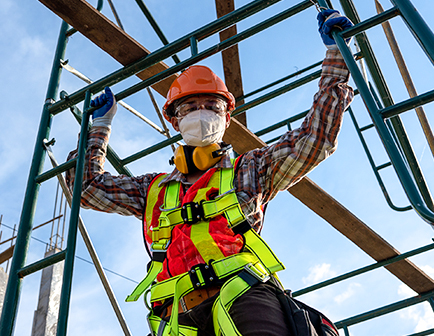 Trabajador de la construcción en un armazón, usando casco naranja, arnés de seguridad, guantes azules y mascarilla, con cielo despejado y estructuras metálicas de fondo.