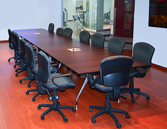Mesa larga de conferencias con sillas giratorias negras, en un entorno de oficina con suelo de madera y paredes claras. Se observan elementos de vidrio al fondo.