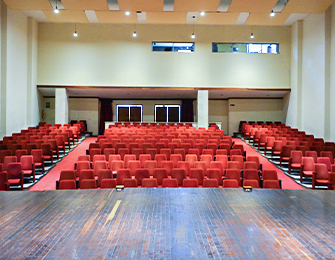 Auditorio con asientos rojos dispuestos en filas, escenario de madera al frente y paredes en tonos neutros, iluminado por luces artificiales y ventanas altas.