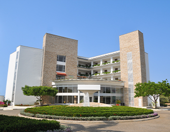 Edificio moderno de varios niveles con fachada de piedra y grandes ventanales, rodeado de áreas verdes y jardines, bajo un cielo despejado.