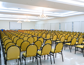 Salón amplio con filas de sillas amarillas dispuestas ordenadamente, iluminación suave y cortinas blancas. Al fondo, una pantalla proyectora está lista para ser utilizada.