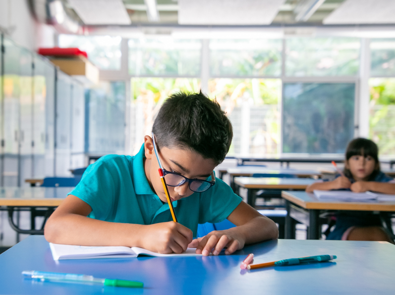 Niño con gafas escribiendo en un cuaderno, rodeado de mesas en un aula iluminada por luz natural.