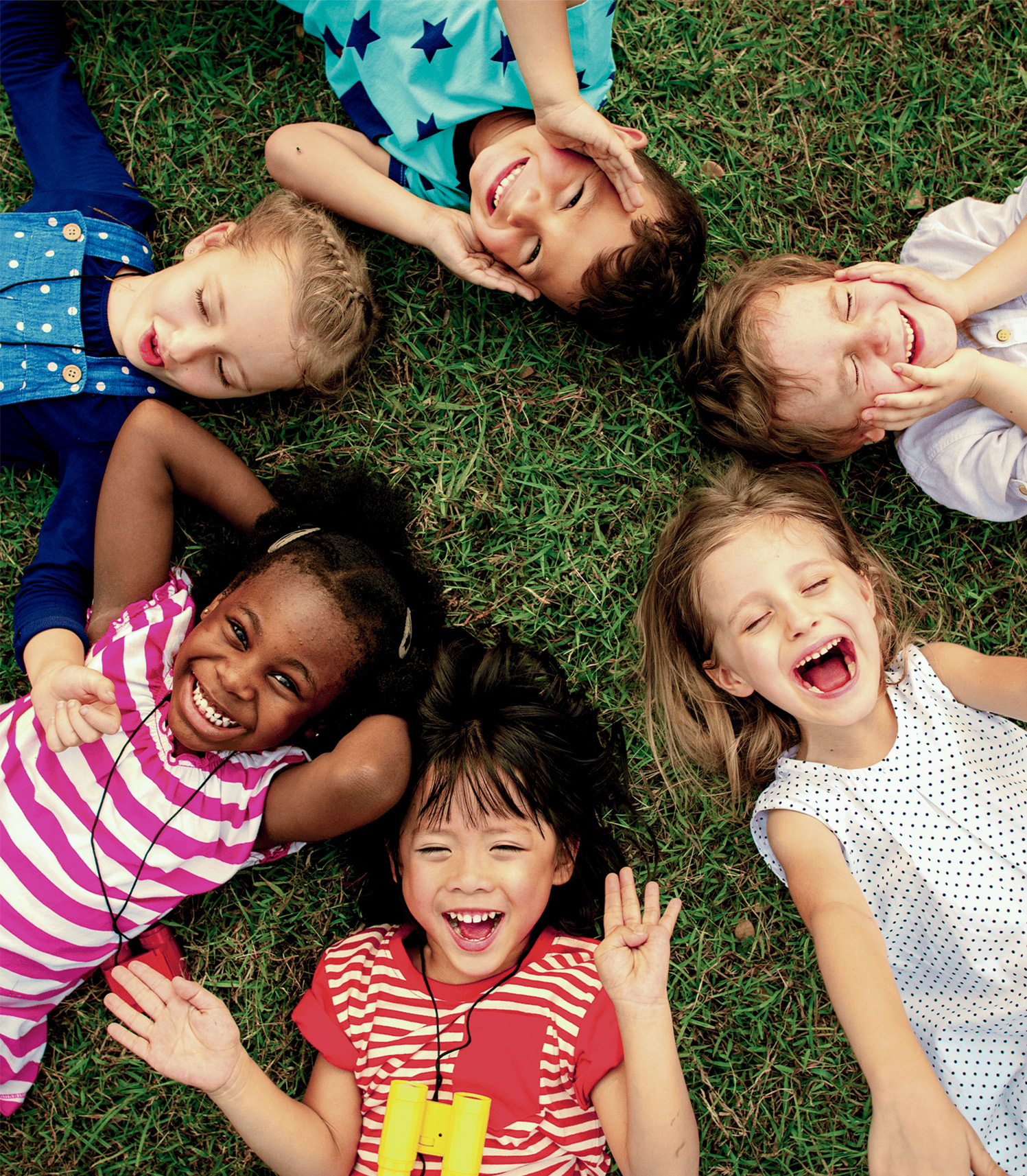 Seis niños sonrientes están acostados sobre el césped, con diferentes camisetas de colores. Algunos posan con las manos en la cara, mientras otros muestran gestos de alegría.
