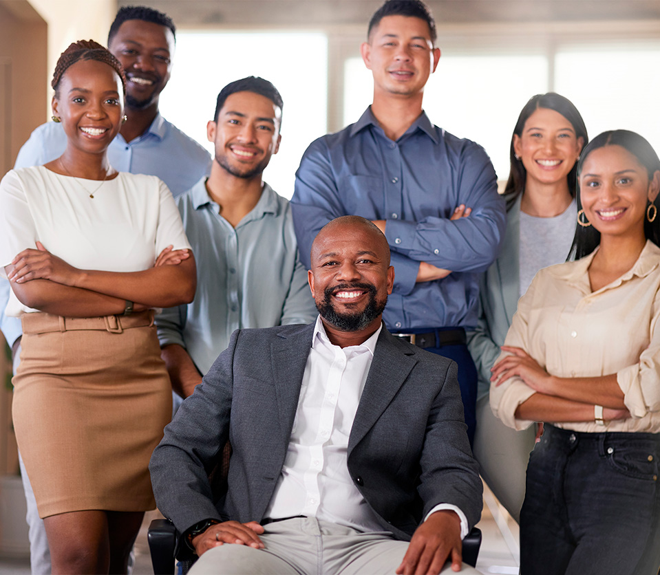 Grupo diverso de seis personas sonriendo, con un hombre sentado al frente. Visten ropa formal y casual, en un entorno iluminado con ventanas y colores neutros.