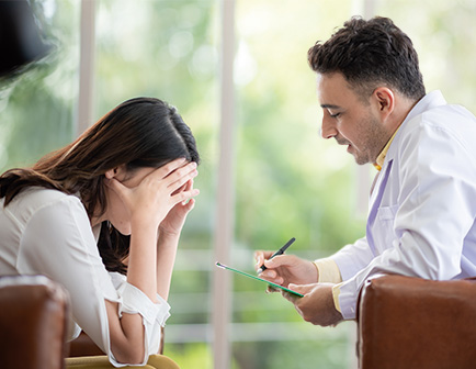 Mujer con las manos en la cabeza escucha a un médico que toma notas en un entorno clínico iluminado.