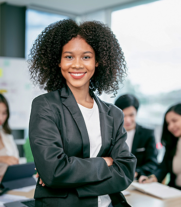 Mujer con cabello rizado y traje negro sonríe frente a un grupo de personas en una oficina moderna. Se observan laptops y documentos en el entorno.