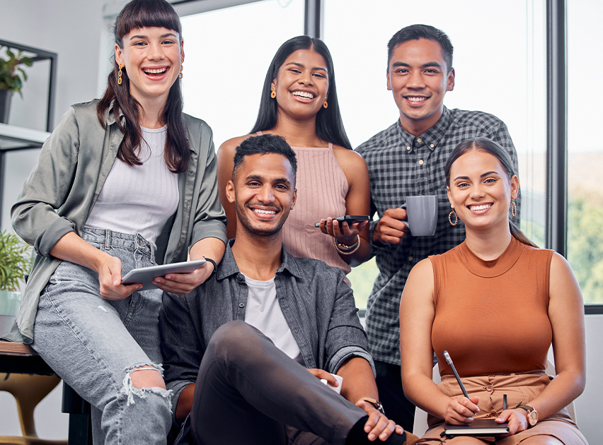 Grupo de cinco personas sonriendo, sentadas y de pie en un entorno luminoso. Sostienen dispositivos móviles y bebidas, vestidas con ropa casual en tonos variados.