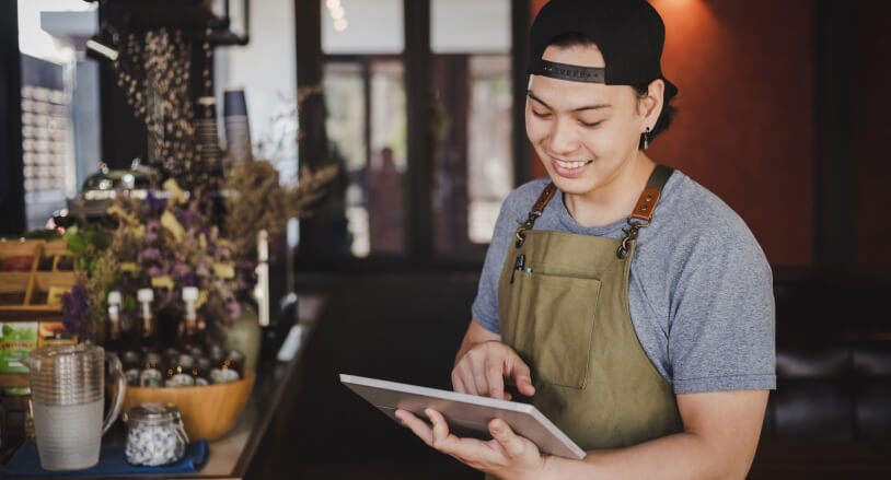 Hombre joven con gorra y delantal, utilizando una tablet en un entorno de café. Fondo con plantas y botellas, iluminación cálida y ambiente acogedor.