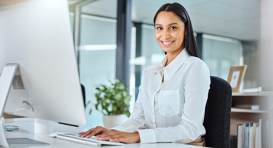 Mujer sonriente sentada frente a un ordenador en un entorno de oficina moderna, con plantas y grandes ventanales que permiten la entrada de luz natural.