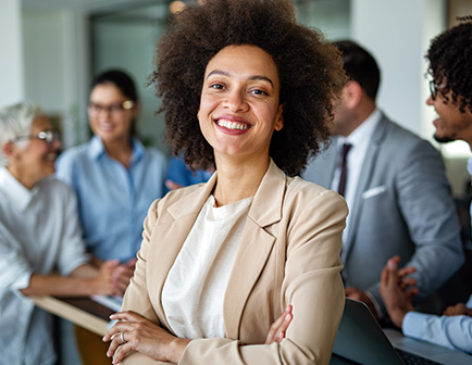 Mujer con cabello rizado y suelto, sonriente, vestida con blazer claro, en un entorno de oficina. Al fondo, varias personas observan, con tonos neutros y un ambiente profesional.