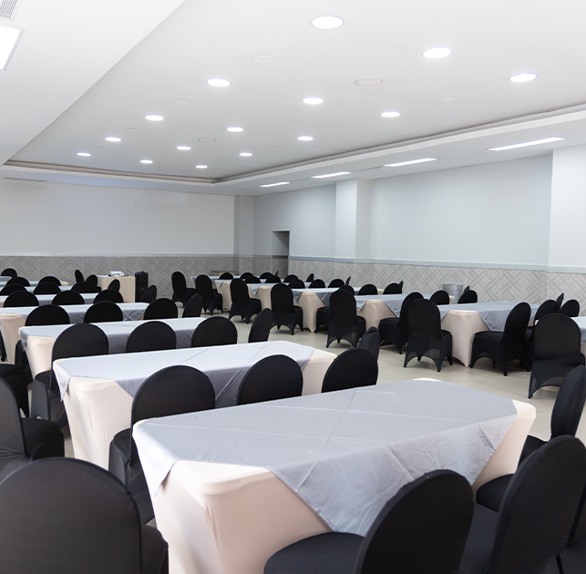 Salón amplio con mesas rectangulares cubiertas de manteles claros y sillas negras, iluminado con luz artificial, paredes en tonos claros y un diseño sencillo.