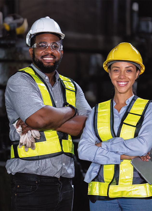 Dos trabajadores sonrientes, uno con gafas y casco amarillo, y otro con casco amarillo y chaleco reflectante, se encuentran en un entorno industrial con maquinaria de fondo.