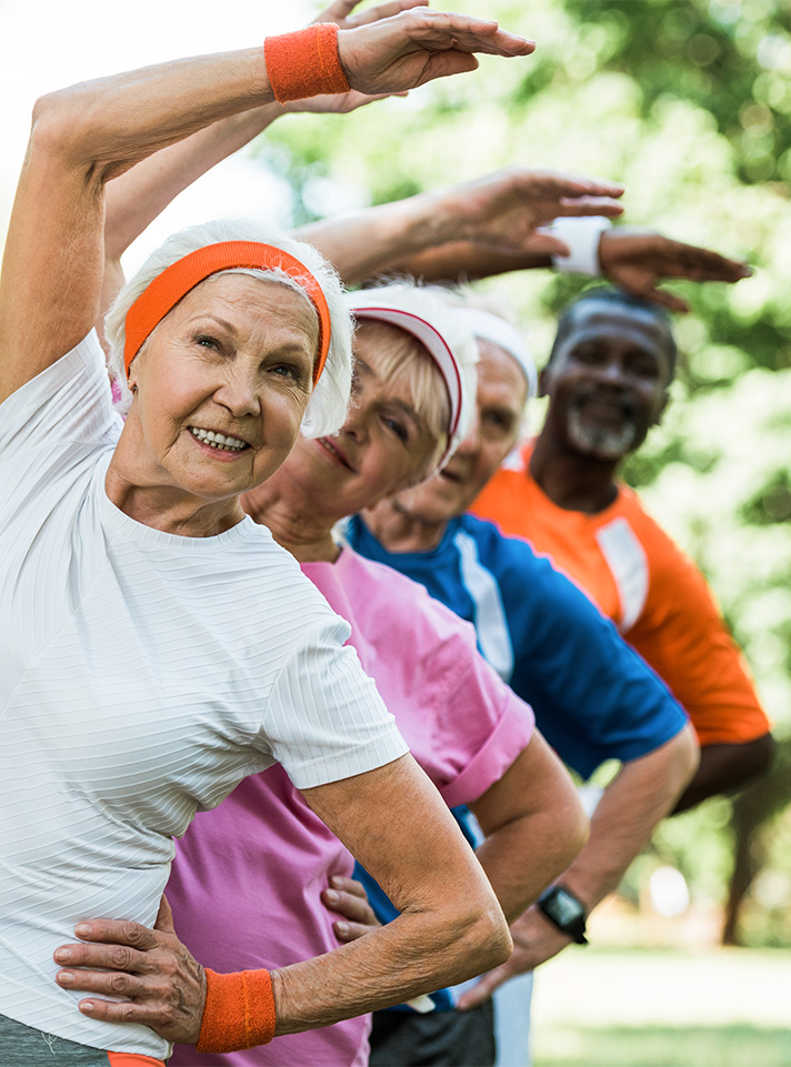 Grupo de personas mayores realizando ejercicios de estiramiento al aire libre, vistiendo ropa deportiva de colores variados, con un ambiente natural y soleado.