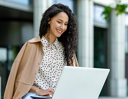 Mujer con cabello rizado y oscuro, sonriente, usando una blusa de lunares y una chaqueta clara, trabajando en una laptop en un entorno exterior con arquitectura moderna.