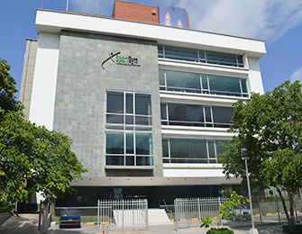 Edificio moderno de varios pisos con fachada de piedra y ventanas grandes, rodeado de árboles y vehículos estacionados, bajo un cielo parcialmente nublado.