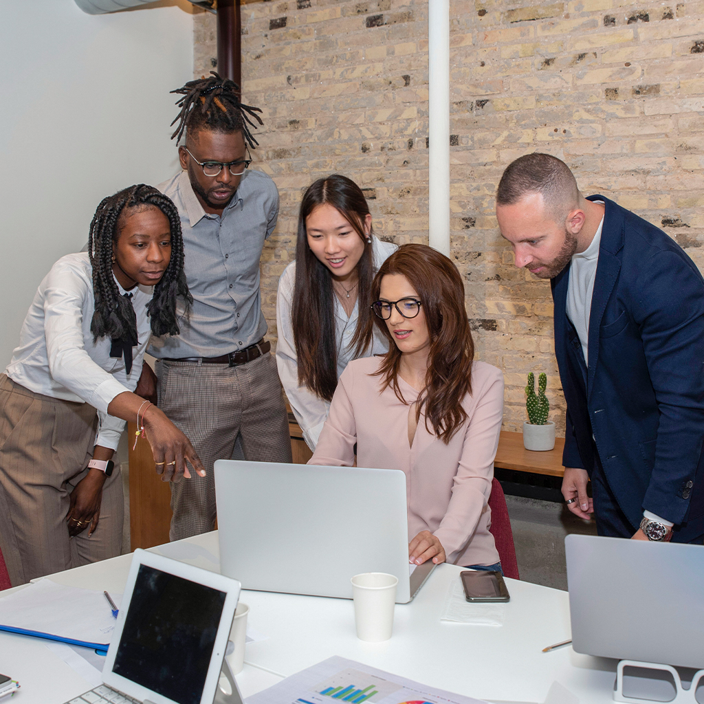 Grupo de cinco personas en una reunión, observando una computadora portátil. Dos hombres y tres mujeres, con un entorno de oficina moderno y paredes de ladrillo expuesto.