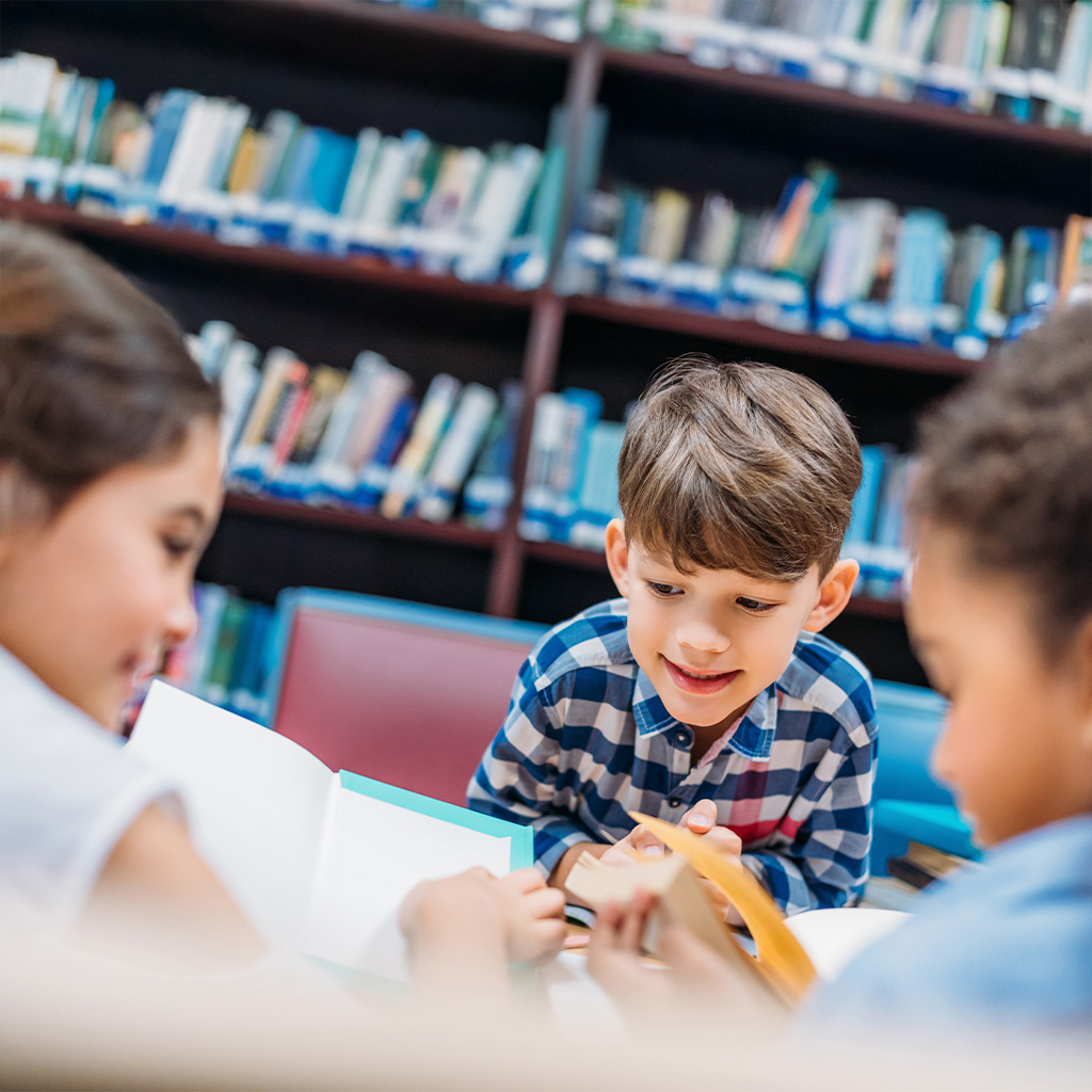 Tres niños interactúan en una mesa en una biblioteca, revisando libros. Al fondo, estantes llenos de libros de diferentes colores. El ambiente es acogedor y educativo.