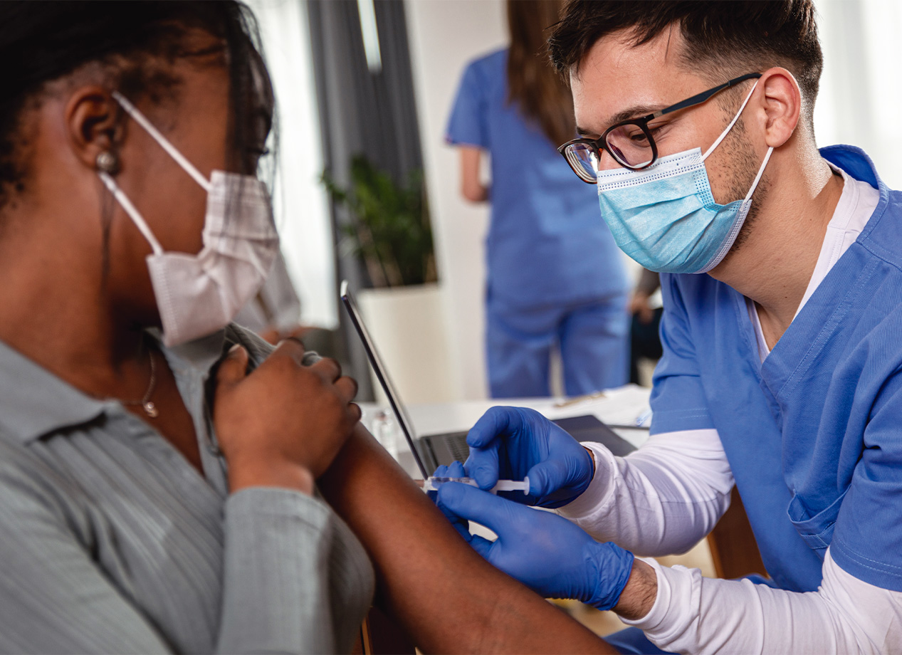 Mujer con mascarilla recibe una inyección en el brazo por un profesional de salud, que también lleva mascarilla. Fondo con otros profesionales y un ambiente clínico.